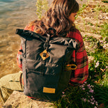 Ein schwarzer JanSport Hatchet Rolltop Adventure Rucksack, getragen von einem Teenager am Rande eines Flussufers.