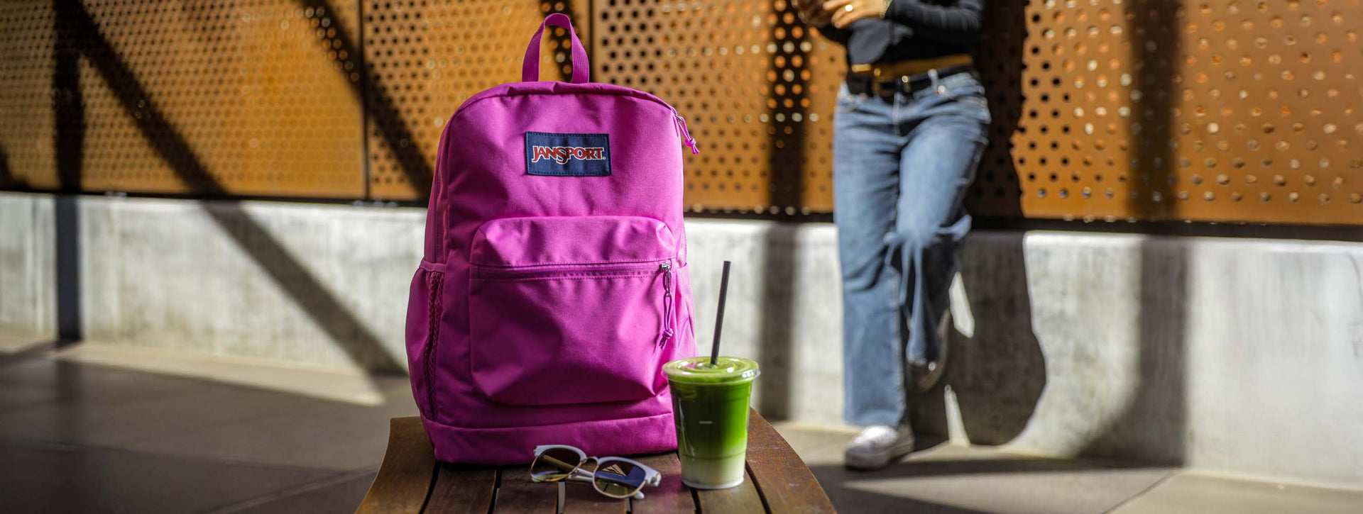 Ein lila Cross Town Plus Rucksack steht auf einem Tisch neben einem Eiskaffee mit Matcha; im Hintergrund steht eine Person. 