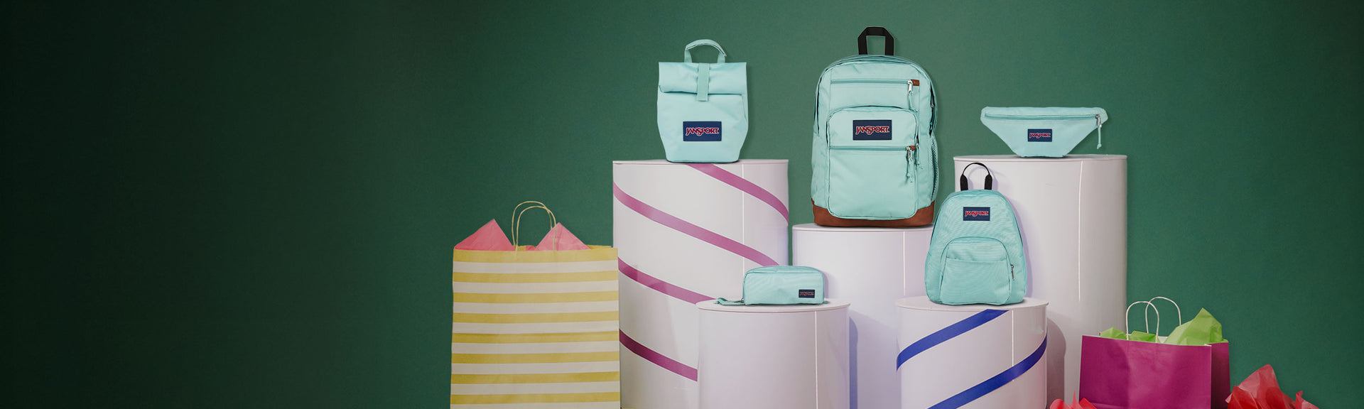 Mint green jansport backpacks on pink cylindrical stands against a green background - jansport cool student, jansport snack attack, jansport waistpack, jansport large accessory pouch, jansport half pint bag. 