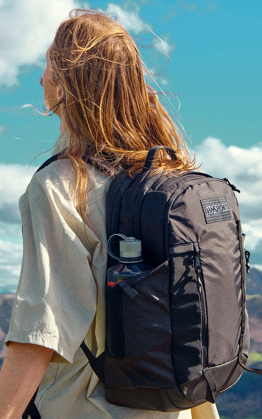Person mit schwarzem Jansport-Rucksack steht auf einem Berg mit malerischer Aussicht