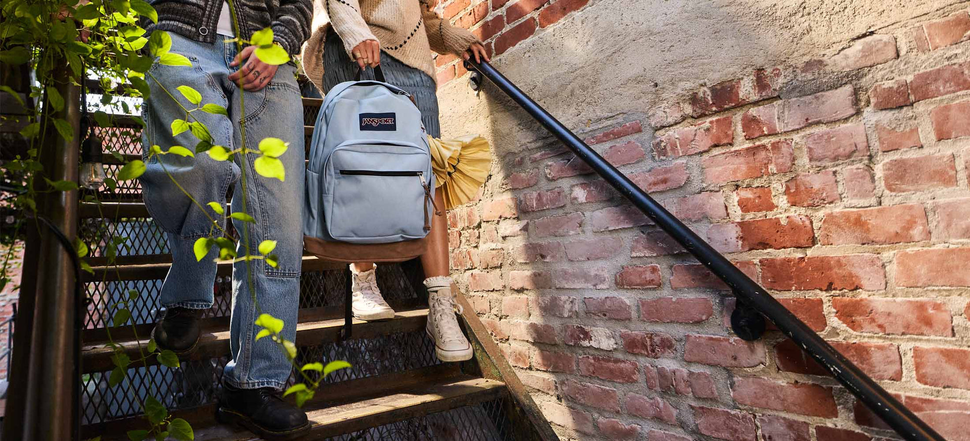 Zwei Personen auf einer Treppe, eine davon trägt einen blauen Jansport-Rucksack, umgeben von Grün und einer Backsteinmauer.